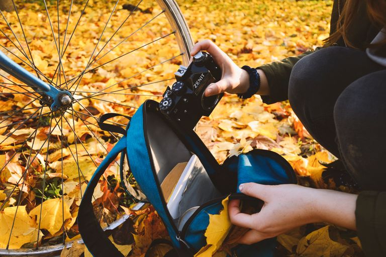 Close-up of individual packing a camera into a backpack next to a bicycle, surrounded by autumn leaves.